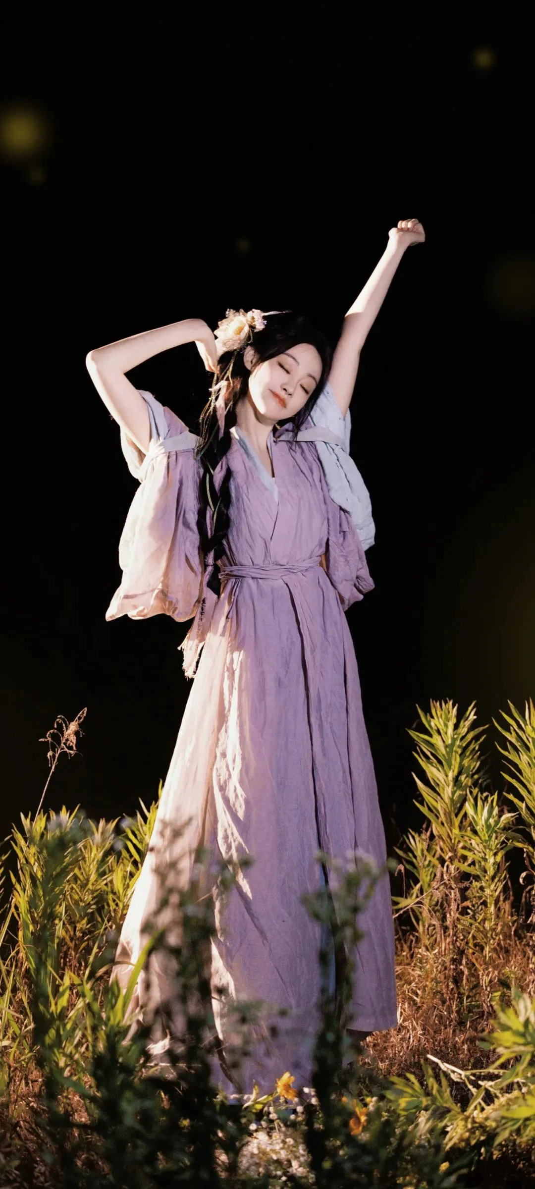 一名身穿淡紫色连衣裙的女子站在夜色中的植物与野花之间。