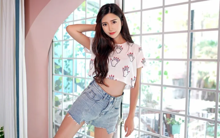 一名长发女性站在明亮的窗边，身穿印花短上衣和牛仔短裤。