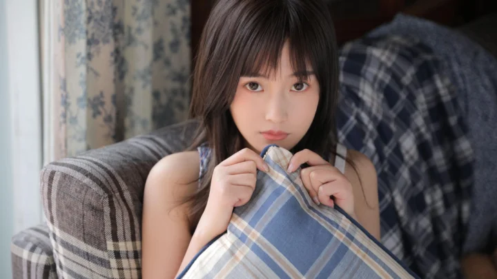 一名女子坐在沙发上，双手抱着一条格纹毯子，窗边光线柔和。