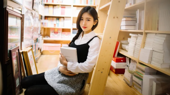 一名成年女性坐在书店书架之间，怀抱一本书。