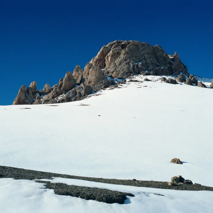 深蓝天空下，一座岩石山峰矗立在广阔的白雪坡地上。