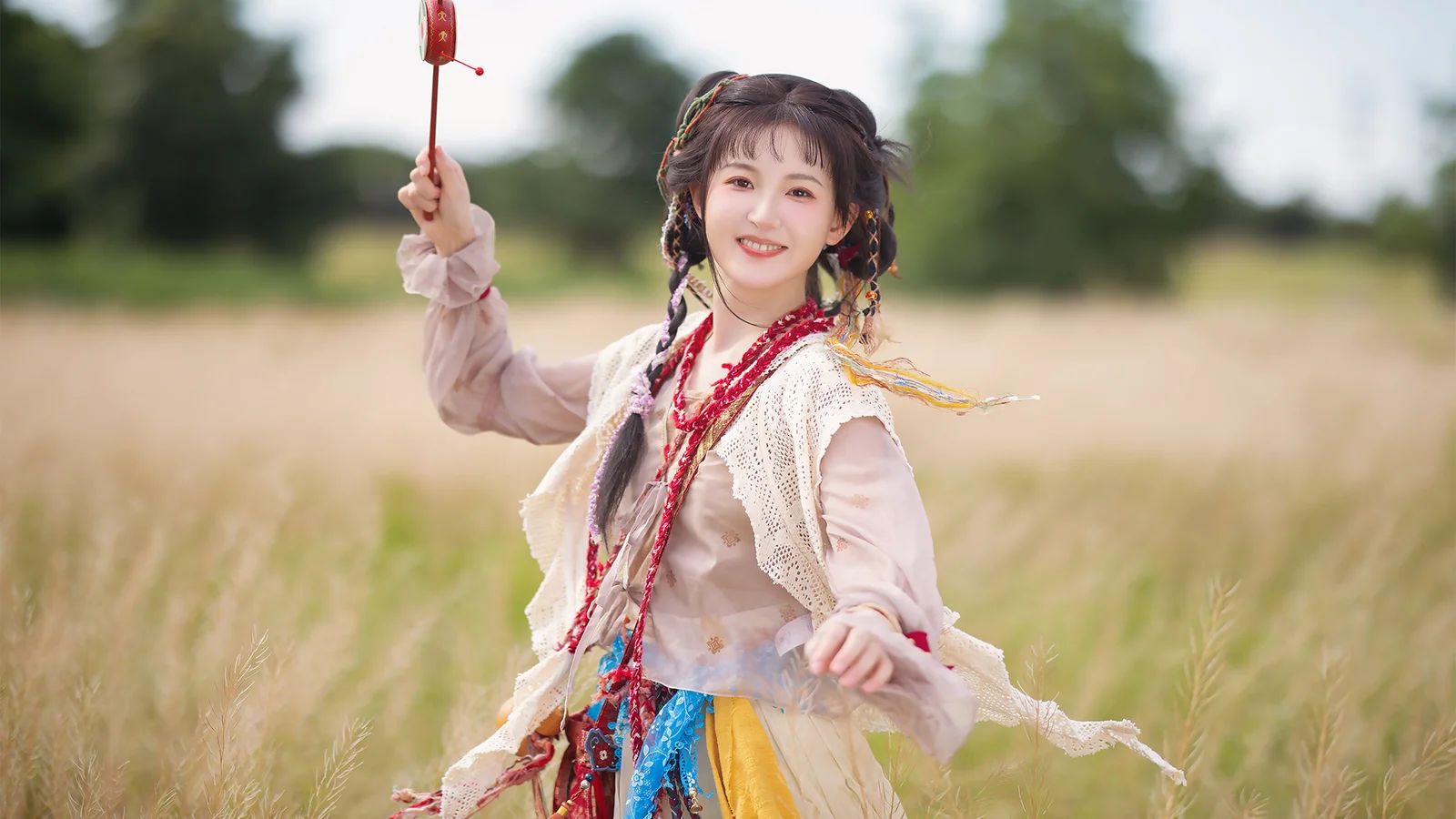 전통 의상을 입은 여성이 들판에 서서 화려한 장식물을 들고 있으며 배경에는 흐릿한 나무와 풀이 보인다.