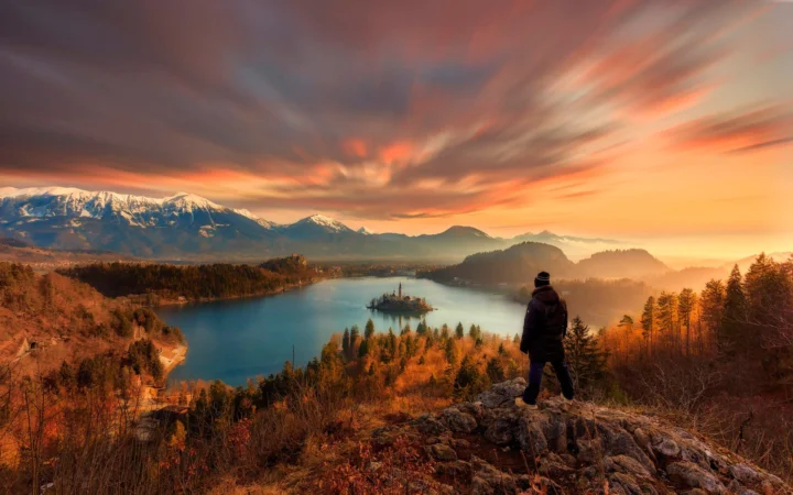 夕阳映照着群山、湖泊与秋色森林，前景岩石上可见一位远处的人。