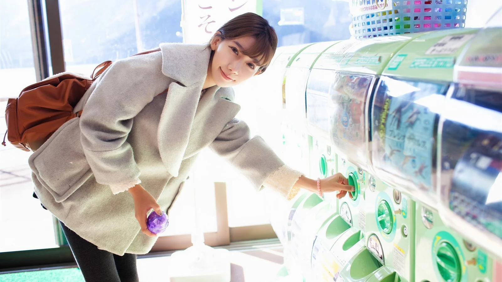 明るい屋内の自動販売機のそばで、淡い色のコートを着た女性が飲み物を取っている。