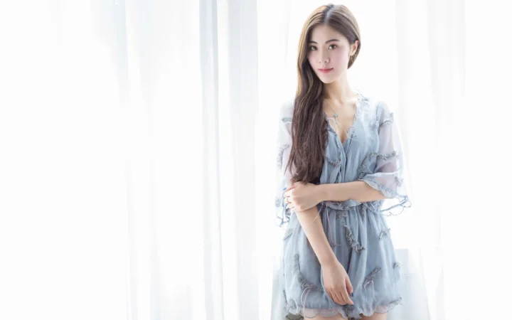 연한 파란색 원피스를 입은 성인 여성이 밝은 커튼 옆에 서 있습니다.