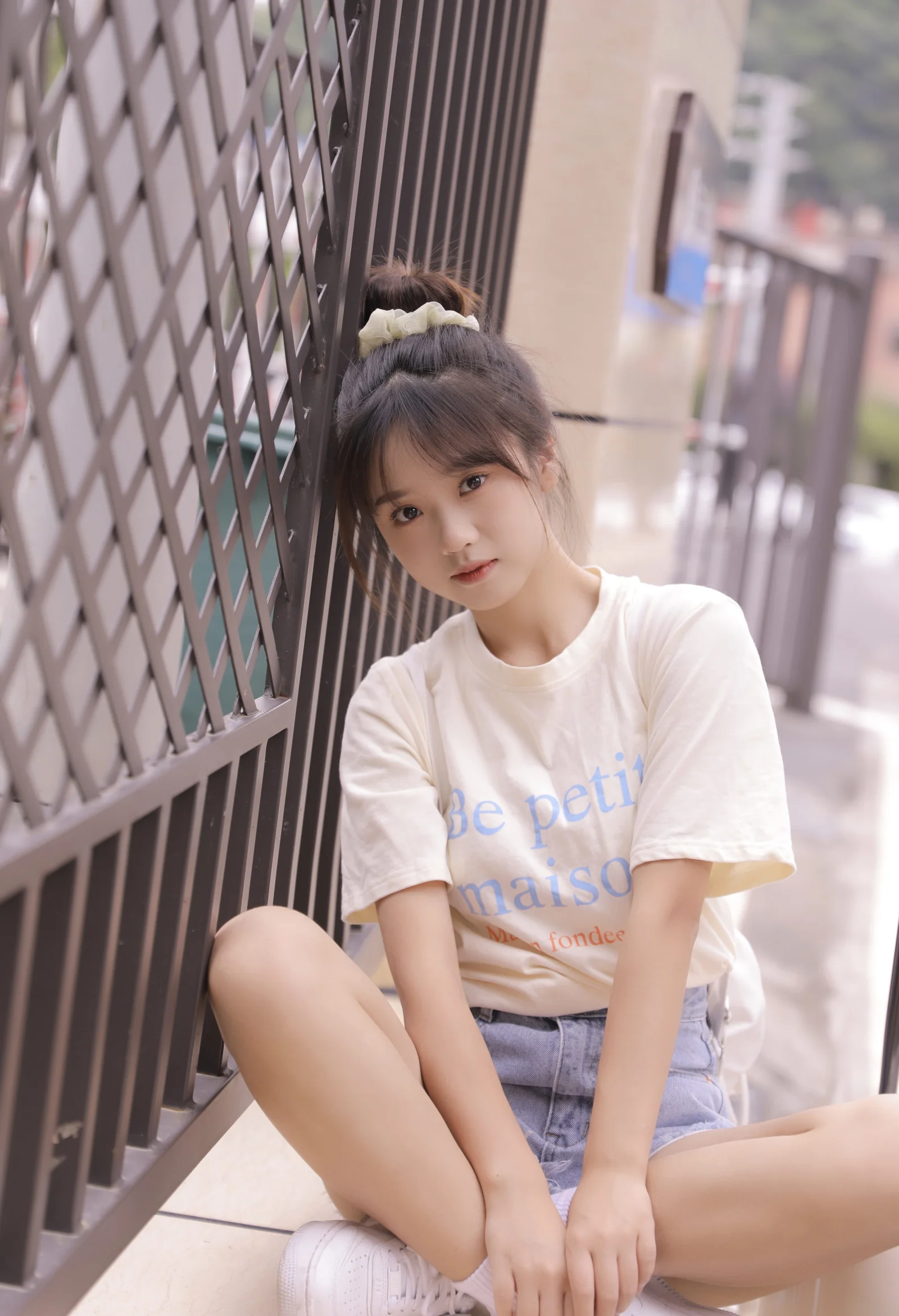 흰색 티셔츠와 데님 반바지를 입은 여성이 금속 울타리 옆에 앉아 있습니다.