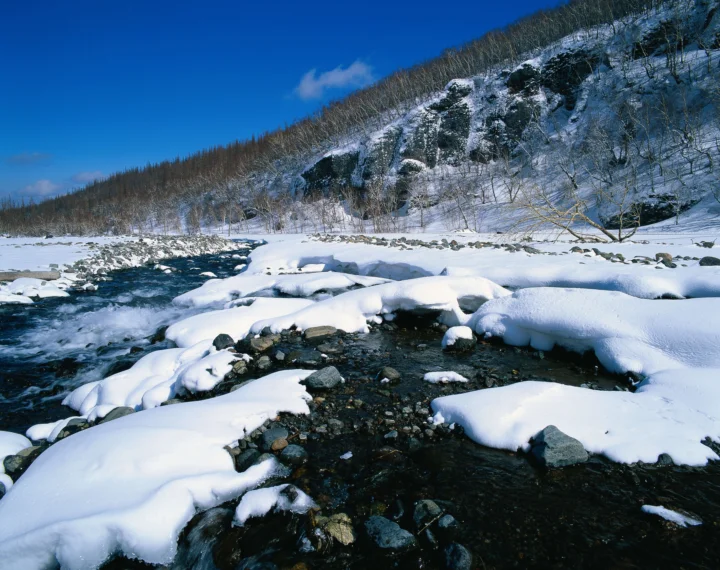 白雪覆盖的山谷中，一条清澈溪流穿过岩石与冰雪，远处是冬日山坡和深蓝天空。