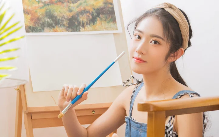 一名女子手持画笔坐在画架前，背景中可见画作。