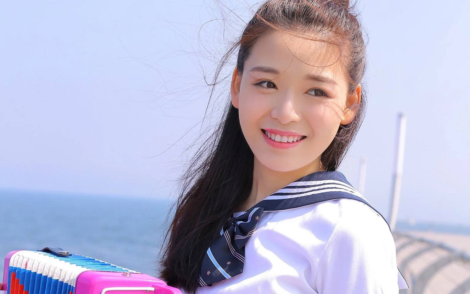 笑顔の成人女性がアコーディオンを持って海辺に立ち、背後に青い海と空が広がっています。