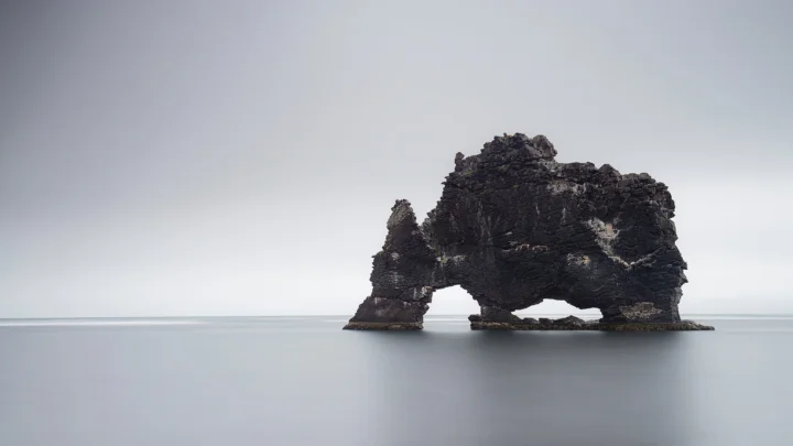 一座深色岩石拱门矗立在平静海面上，远处是朦胧的灰色天空。