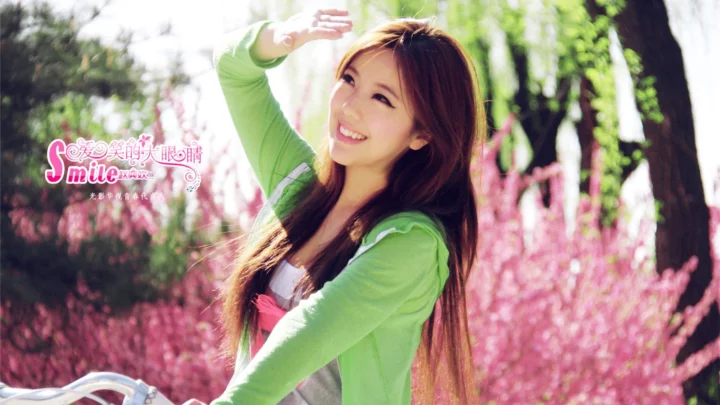 긴 머리의 여성이 초록색 재킷을 입고 분홍색 꽃나무 사이에서 미소 지으며 한 손을 들고 있습니다.