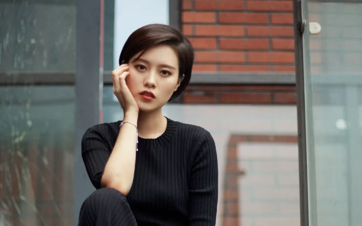 一名成年女子身穿黑色针织衫，坐在砖墙与玻璃门旁看向镜头。