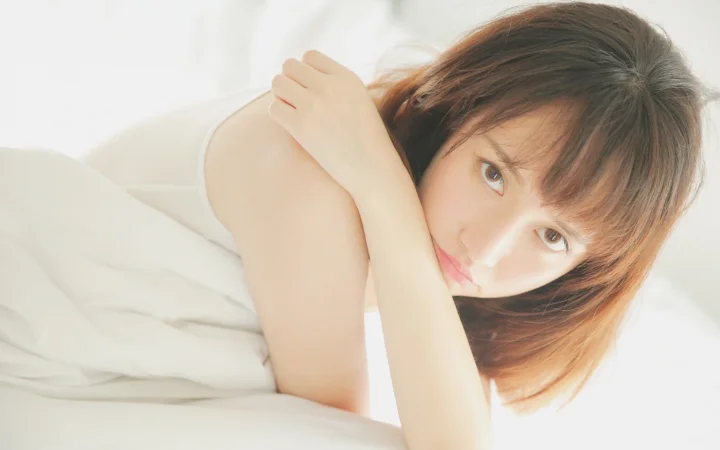 一位棕发女性倚靠在白色床品上，注视着镜头。