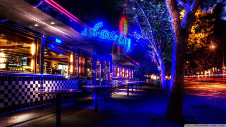 夜晚的城市街道旁，一家餐馆在蓝紫色霓虹灯和树木映衬下熠熠生辉。
