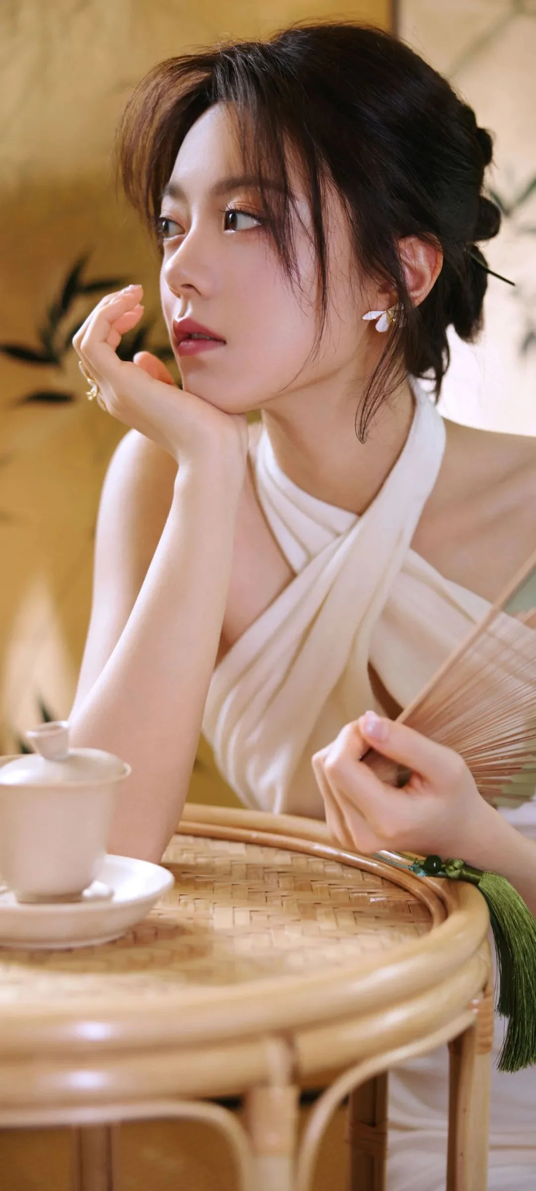 一位女性在暖色室内光线下托腮坐在桌边，手中拿着折扇。
