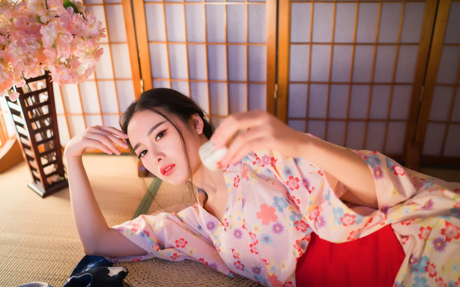 一名成年女子躺在榻榻米房间内，身穿浅色花卉和服风格服装，背景是木格移门与粉色花枝。