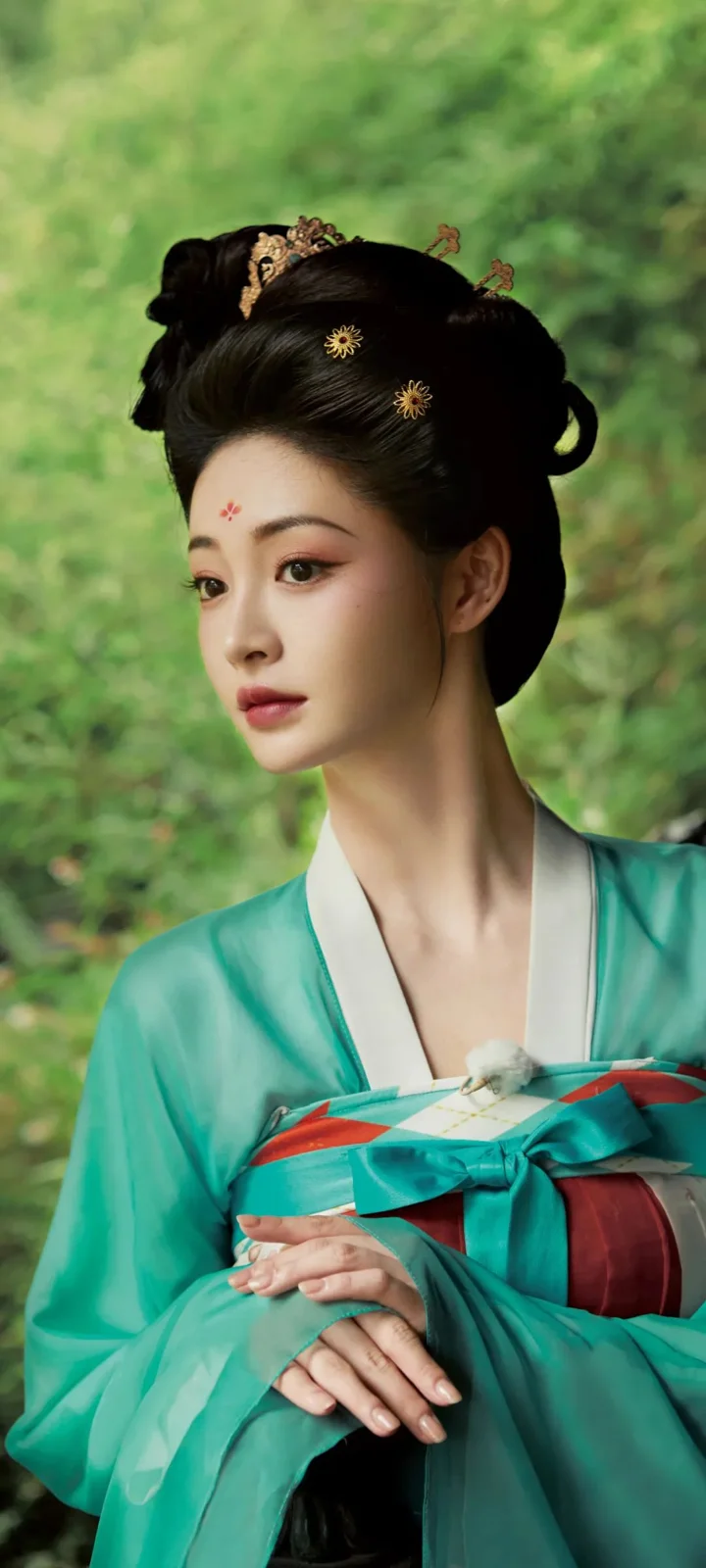 一名成年女子身穿青绿色传统服饰，在绿色户外背景前侧身凝视。