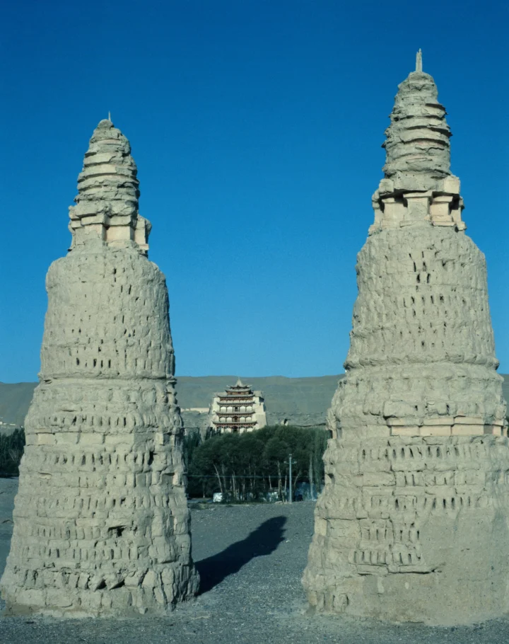 两座高大的石塔矗立在前景，中间远处可见一座多层古建筑，背景是蓝天与山丘。