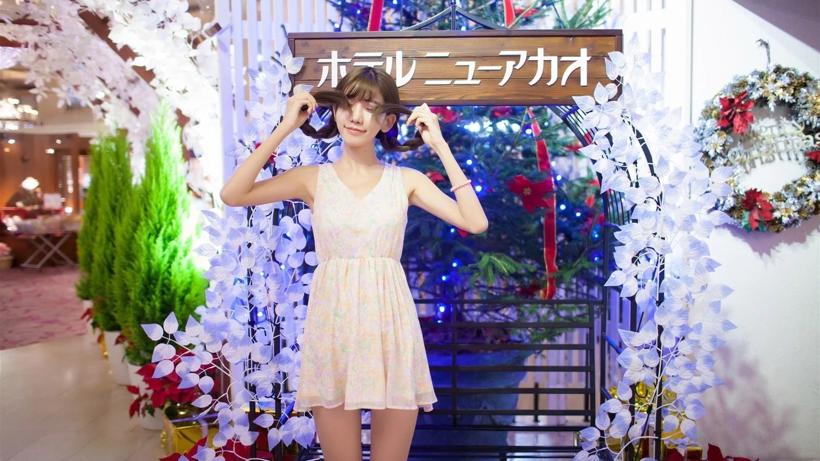 女性が屋内の花飾りのアーチの前で、両手で帽子のつばに触れている。