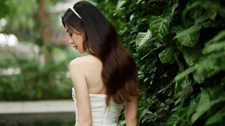 一名长发女子穿着白色裙装站在茂密的绿色植物旁。