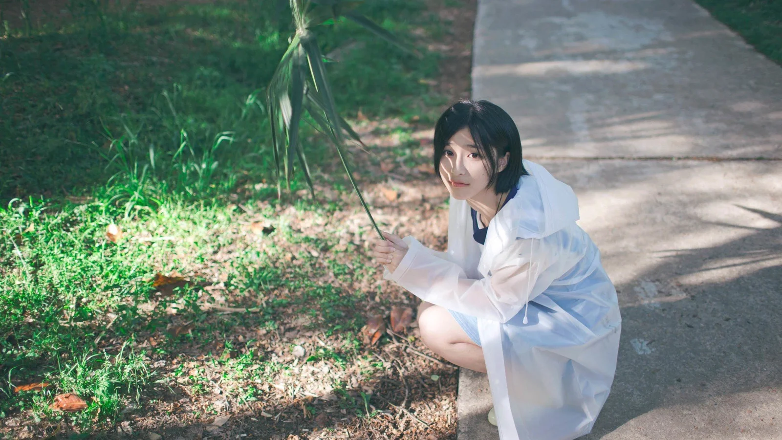 透明なレインコートを着た女性が、木陰の小道のそばにしゃがんでいる。
