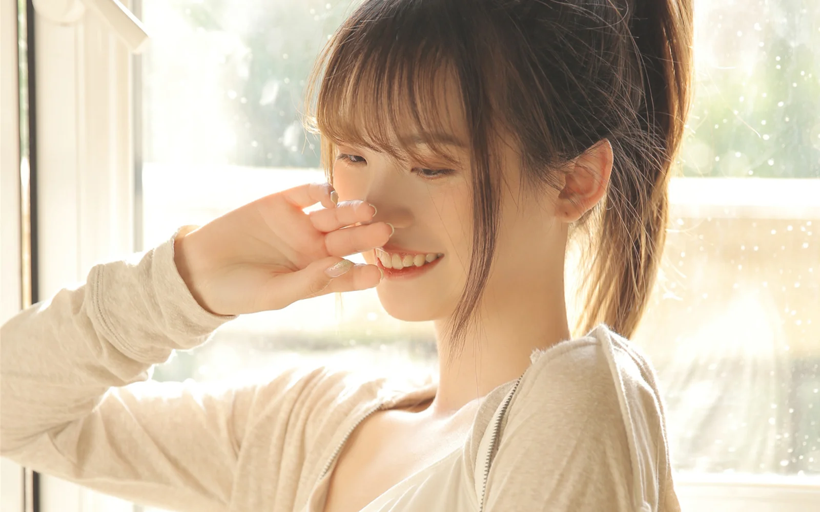 창가의 부드러운 자연광 속에서 성인 여성이 입가에 손을 대고 미소 짓고 있다.