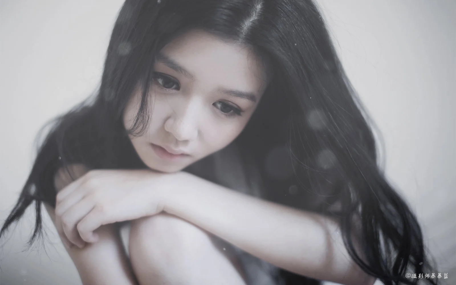 一名黑发女性低头靠在手臂上，画面采用柔和的灰白色调。