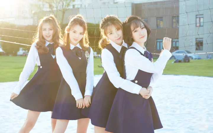 어두운 색상의 교복을 입은 네 명의 여성이 야외 배구 네트 앞에 서서 뒤쪽에서 비치는 햇빛을 받고 있습니다.