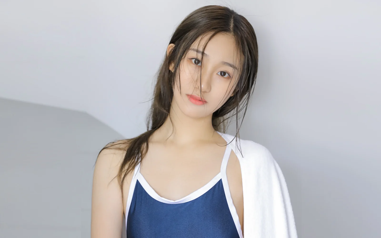 一名长发女子身穿蓝色吊带上衣并披着白色外套，在浅灰色背景前看向镜头。