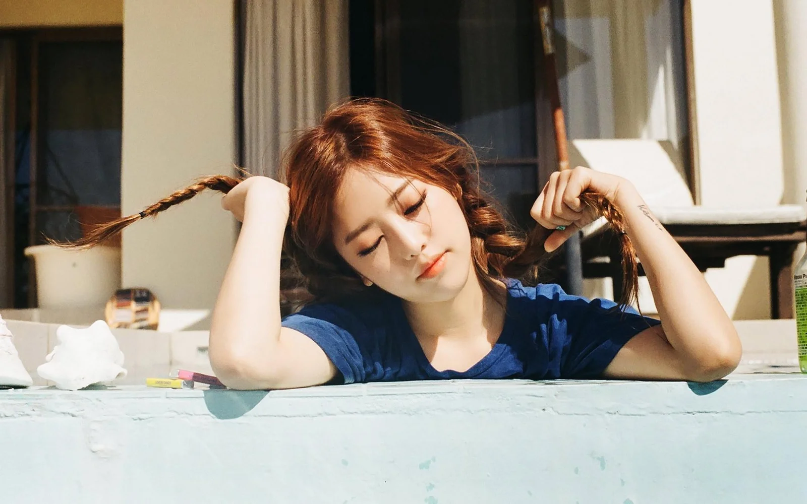 一名棕发女子穿着蓝色上衣，双手轻握辫子，倚在窗台上沐浴阳光。