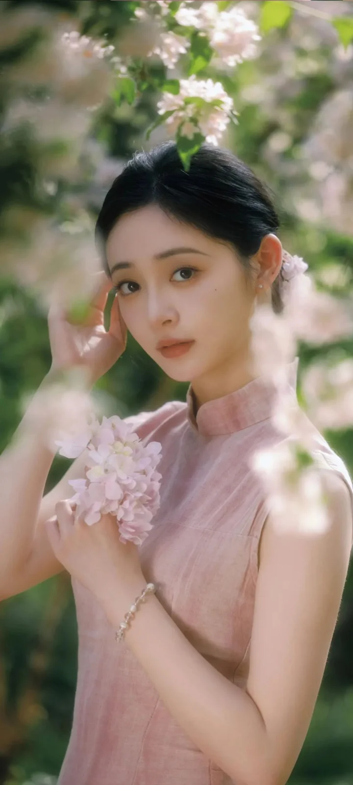 ピンクの服を着た女性が、花の咲く枝と柔らかな緑の中で淡い色の花束を持っています。