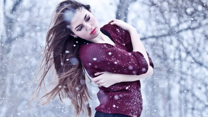一名成年女子穿着酒红色针织衫站在飘雪的冬季树林中。