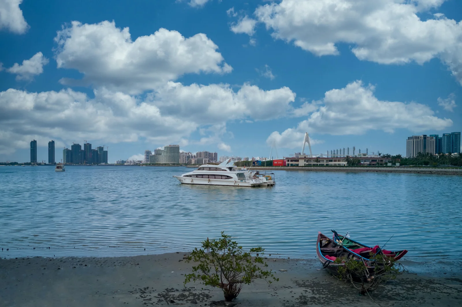 蓝天下的海湾水面前方是一片城市天际线，水面上有船只，岸边还有一艘小渔船。
