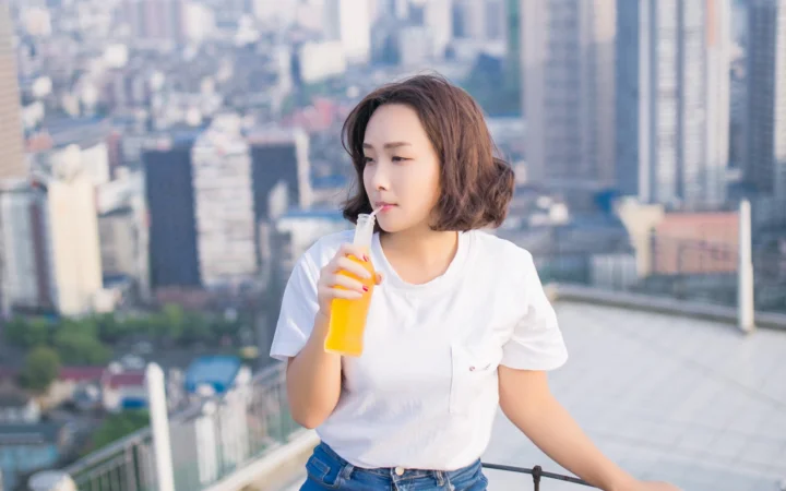 一名成年女性在城市天台上手持黄色饮料，身后是虚化的高楼建筑。