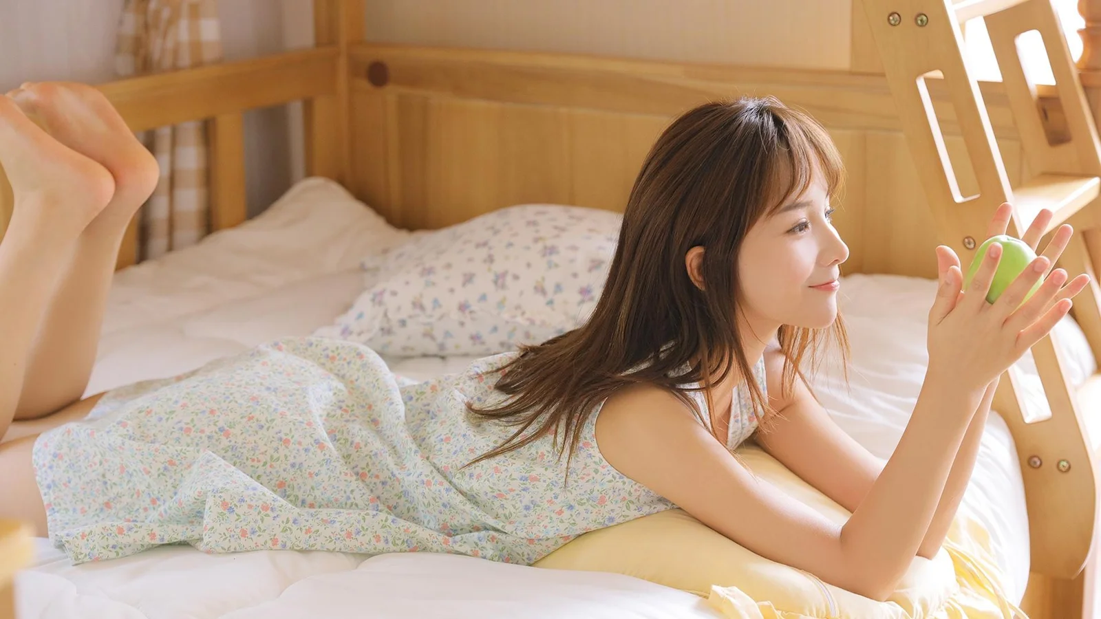 一名女子趴在阳光明亮的卧室床上，手中拿着一个绿色物体。