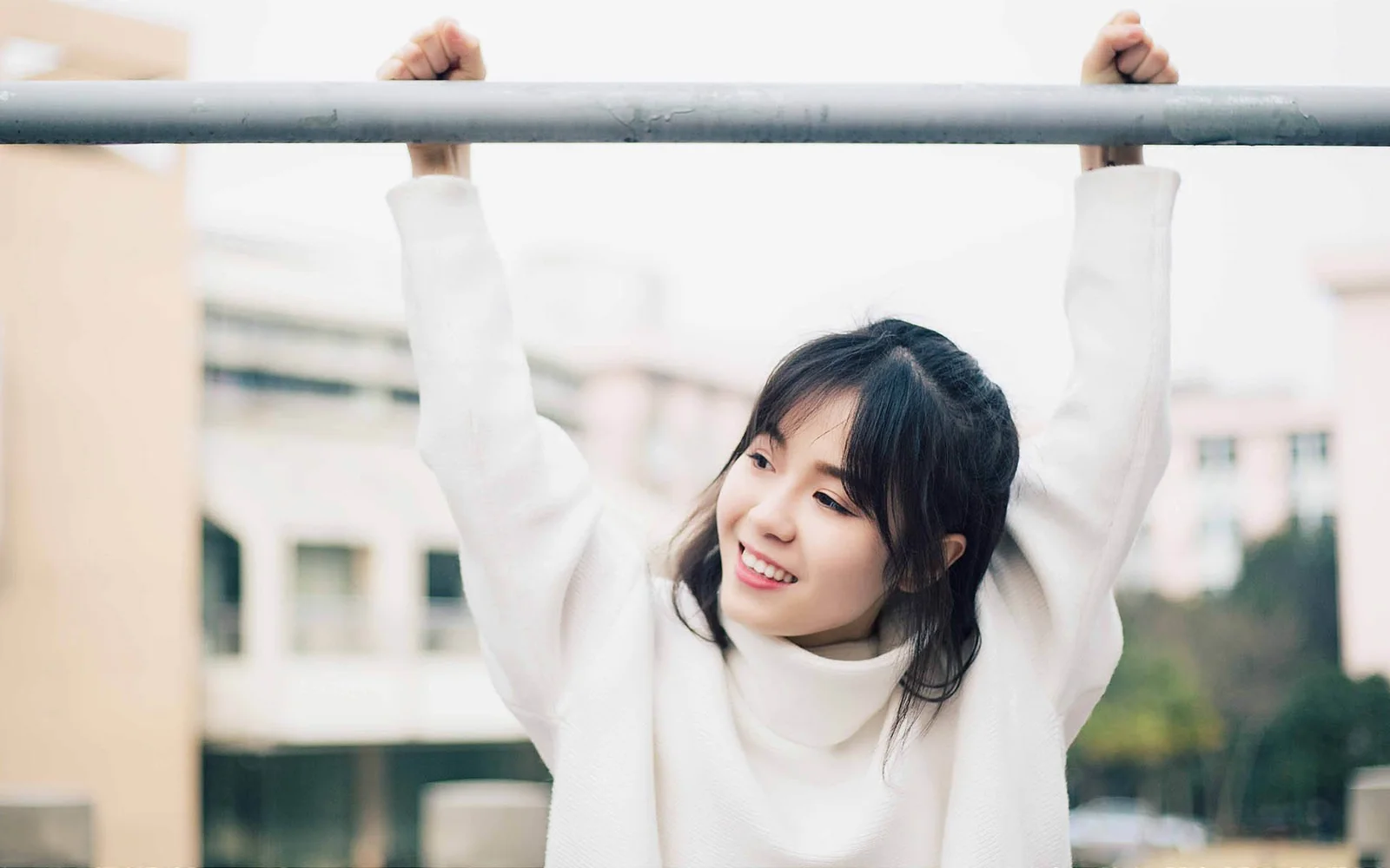 흰색 스웨터를 입은 여성이 야외에서 가로봉을 잡고 있으며 배경에는 흐릿한 건물이 보입니다.