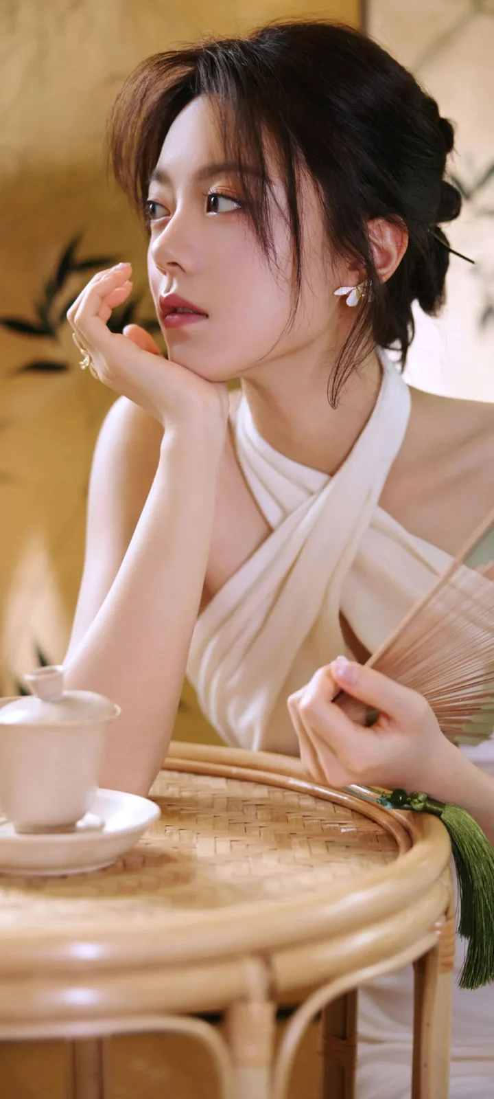 一位女性在暖色室内光线下托腮坐在桌边，手中拿着折扇。