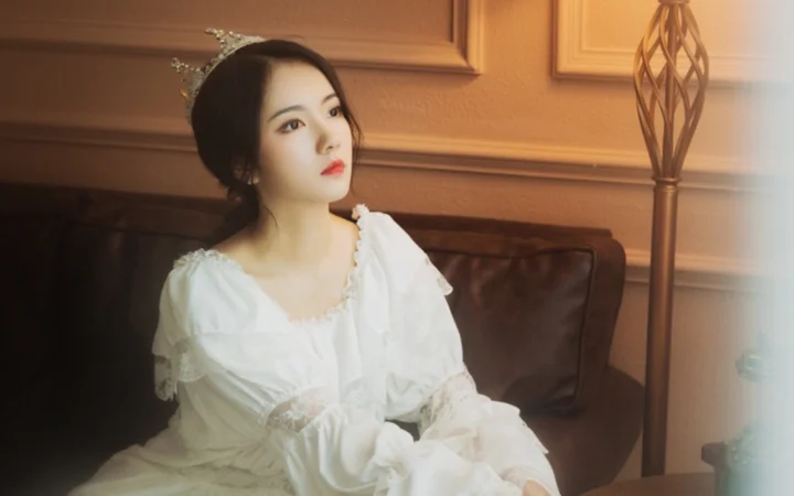 티아라를 쓰고 흰 드레스를 입은 여성이 실내 소파에 앉아 있다.