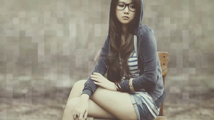 一名戴眼镜的长发女性坐在椅子上，身后是柔和的方格纹理背景。