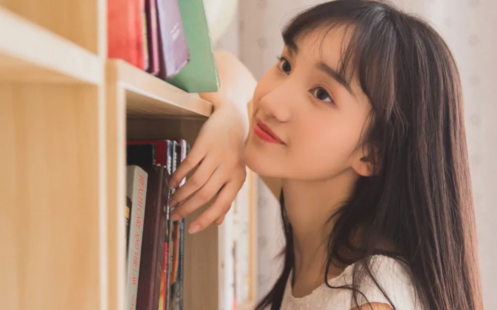 一名长发女子靠在书架旁，面向镜头微笑。
