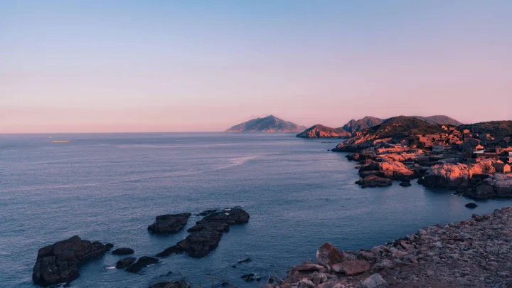 晨雾中的海岸、礁石、海面与远处岛屿沐浴在粉紫色的日出光线中。