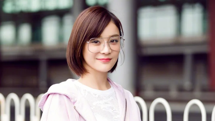 一位戴眼镜的短发女性身穿浅粉色外套，站在虚化的建筑背景前。