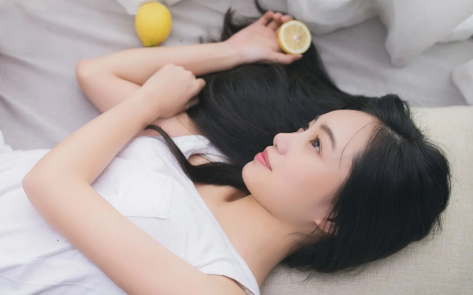 一名成年女子身穿白色上衣躺在床上，手持半片柠檬望向一侧。