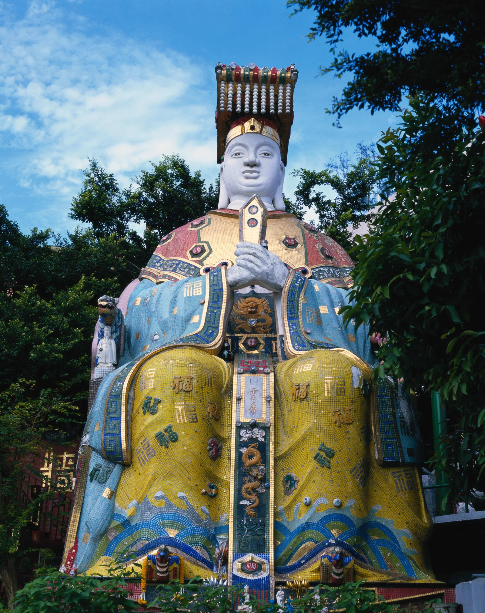 青空の下、色鮮やかな装飾衣装をまとった大きな像が木々に囲まれて立っています。