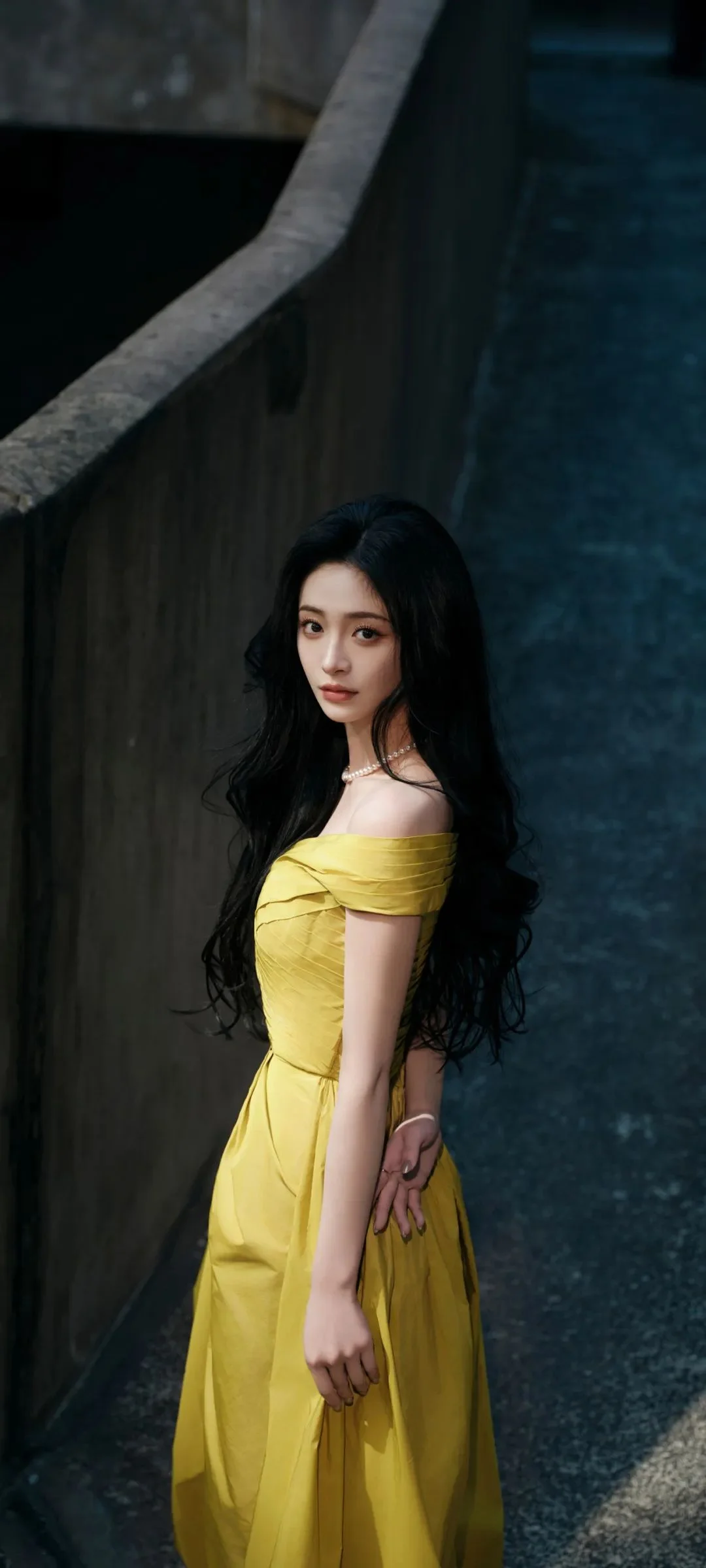긴 머리의 여성이 노란 드레스를 입고 어두운 콘크리트 통로 옆에 서 있습니다.
