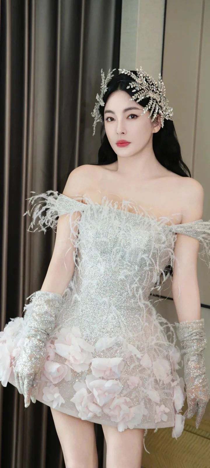 一名成年女性身穿白色花饰礼服，佩戴华丽头饰，站在深色窗帘前。