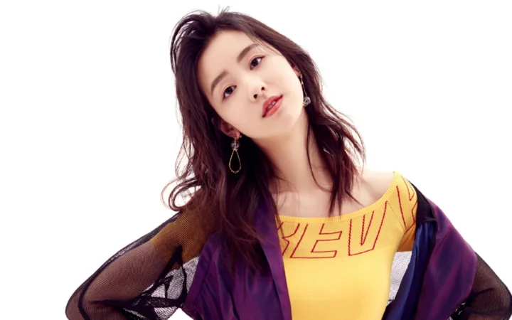 一名成年女性穿着彩色服装，在白色背景前侧头面对镜头。