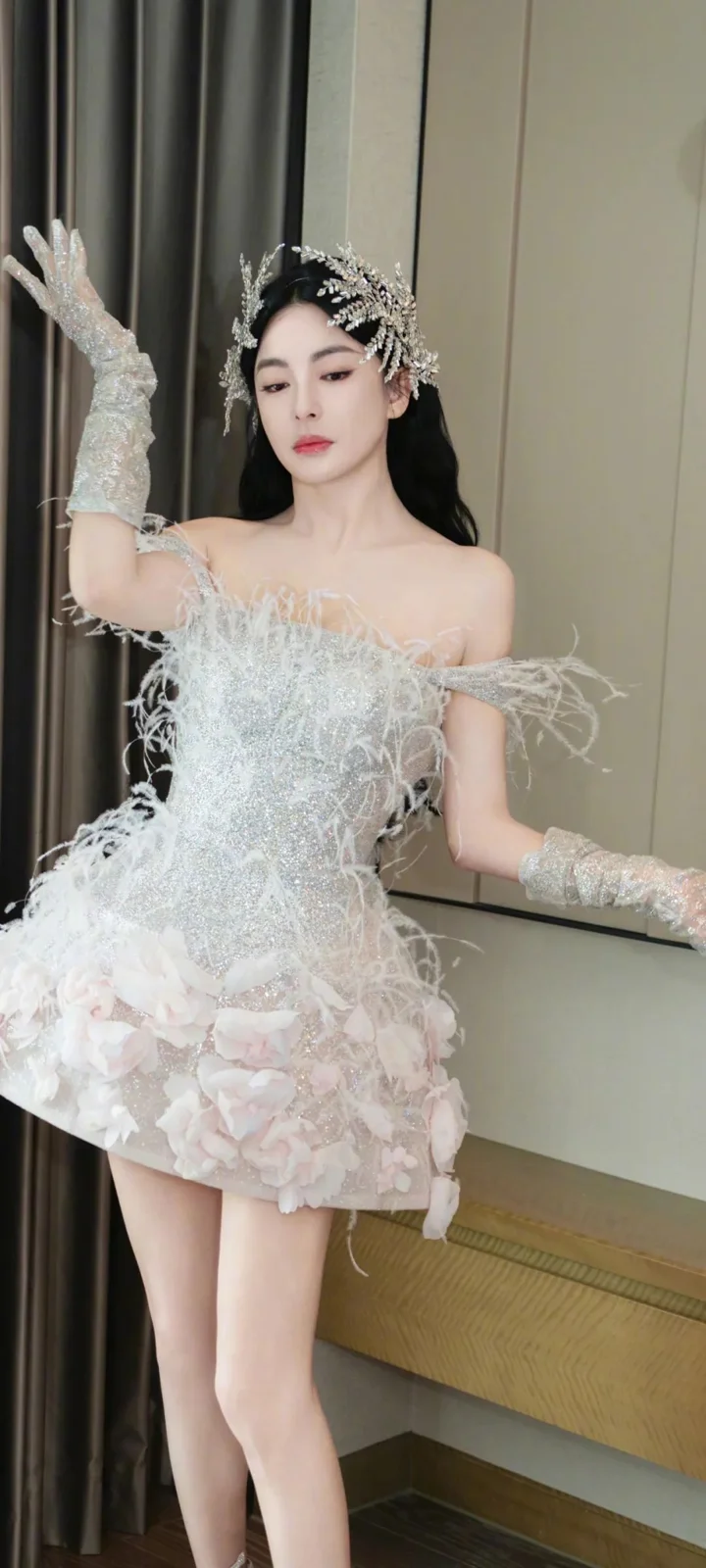 一位成年女性身穿白色花朵装饰礼服，佩戴闪亮头饰，站在室内窗帘与墙面前。