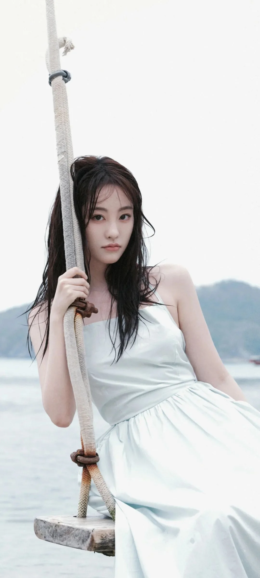 一名女子穿着白色连衣裙坐在海边秋千上，背景是海水与远山。