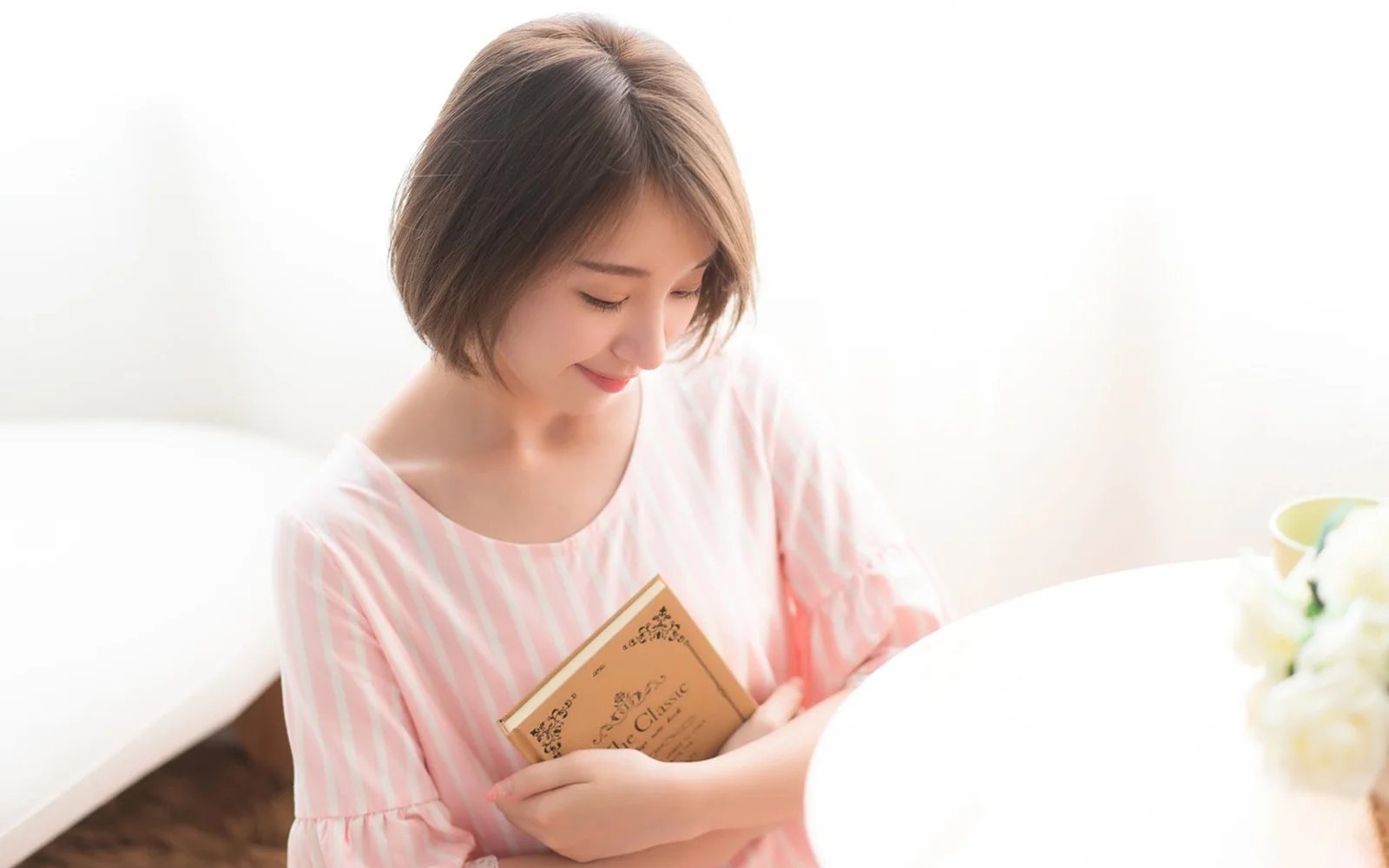 분홍색 줄무늬 상의를 입은 성인 여성이 흰색 테이블 옆에서 책을 읽고 있다.